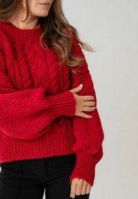 Sarkanais adīts džemperis ar kabelveida rakstu, ribotām piedurknēm un brīvu piegriezumu. Izgatavots no bieza, ērta materiāla.