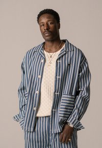 Camicia-giacca a righe blu e bianche con grandi bottoni, dotata di una tasca sul petto e un design unico delle maniche. Indossata sopra un top crema testurizzato.