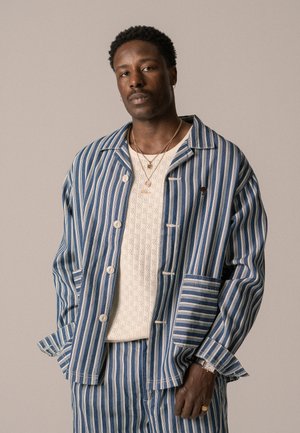 Chemise-veste rayée bleu et blanc avec de grands boutons, comportant une poche poitrine et un design de manches unique. Portée par-dessus un haut crème texturé.