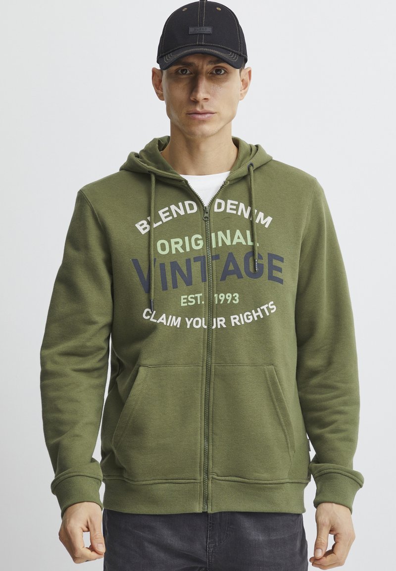 Sudadera verde con cremallera hecha de un tejido suave, con un estampado de estilo vintage en blanco y marino, con un bolsillo canguro y capucha ajustable.