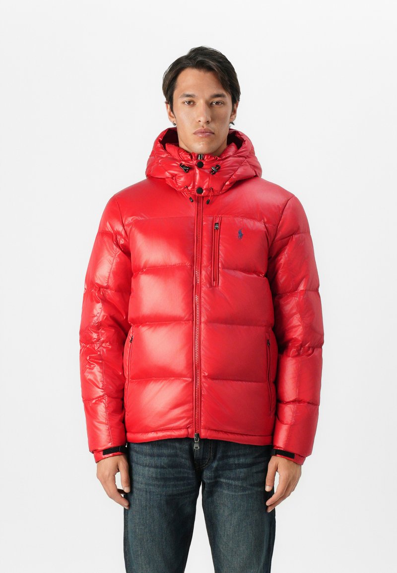 Mann, der eine leuchtend rote Steppjacke mit Kapuze und Reißverschlusstaschen trägt, vor einem schlichten weißen Hintergrund steht.