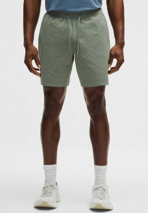 Mann trägt hellgrüne Shorts mit Kordelzug, weiße Bündchensocken und weiße Sportschuhe und steht vor einem einfarbigen Hintergrund.