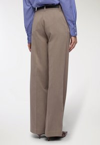 Kobieta stojąca, ubrana w beżowe szerokie spodnie, niebieską i białą w paski koszulę oraz brązowe buty na obcasie, pokazana z tyłu.