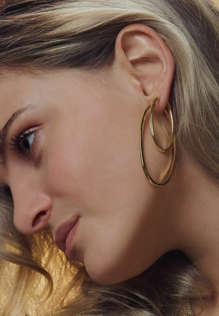 Guldörhängen i en lagerdesign med en glänsande finish, som visar upp släta kurvor och ett lätt utseende mot texturerat hår.