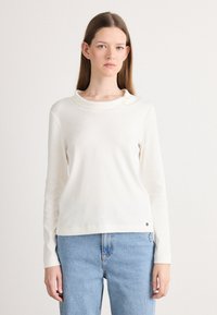 Off-white longsleeve shirt met een kraag, zacht materiaal, relaxte pasvorm en een kleine logo-accent bij de onderrand. Geregardeerd met lichtblauwe jeans.