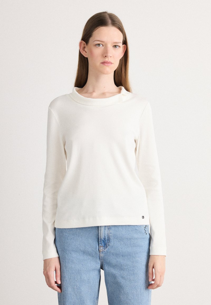 Off-white longsleeve shirt met een kraag, zacht materiaal, relaxte pasvorm en een kleine logo-accent bij de onderrand. Geregardeerd met lichtblauwe jeans.