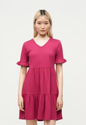 Jeune femme aux cheveux blonds raides portant une robe rose vif à manches courtes, à volants aux manches.