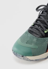 Sneaker verde a rete con dettagli neri, sovrapposizioni testurizzate e suola in gomma con spruzzi. Comprende lacci neri e dettagli riflettenti.