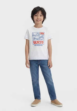 Jongen staat, draagt een wit T-shirt met print "GUESS Los Angeles 1981", blauwe spijkerbroek en beige slip-ons op een witte achtergrond.