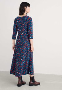 Kukkakuvioinen midi-mekko, jossa on sininen tausta, punaisia ja valkoisia kukkia, kolme neljäsosaa hihat ja kelluva siluetti, yhdistettynä ruskeisiin nilkkabootseihin.