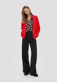 Roter Strickcardigan mit offenem Vorderteil, getragen über einer schwarz gemusterten Bluse und einer schwarzen weit geschnittenen Hose. Schwarze klobige Schuhe runden das Outfit ab.