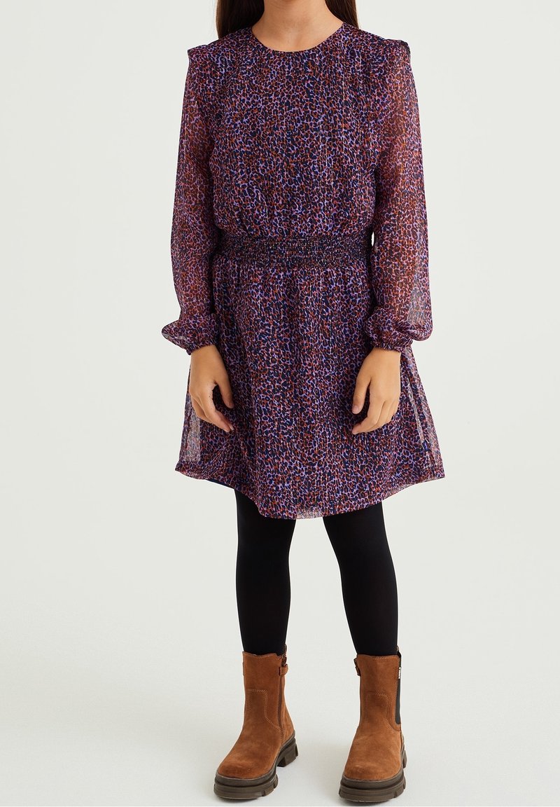 Fille portant une robe à manches longues à imprimé animal violet et marron, des leggings noirs et des bottines en daim marron, debout devant un fond uni.