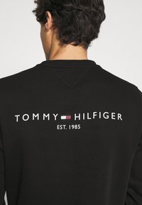 Felpa nera con scollo rotondo e logo sul retro con la scritta "TOMMY HILFIGER" e "EST. 1985", che presenta un design minimalista.