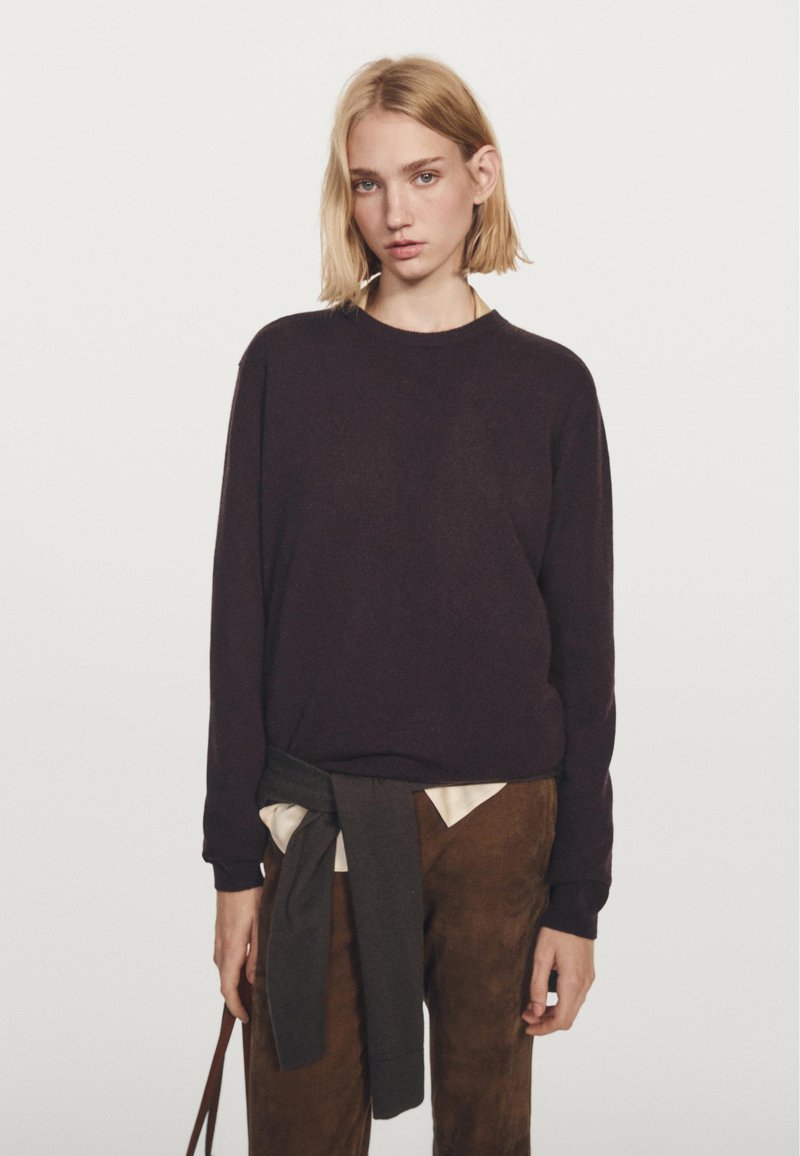 Dark purple sweater with long sleeves and a round neckline, paired with light-colored shirt and brown pants. Sweater has a soft texture.
