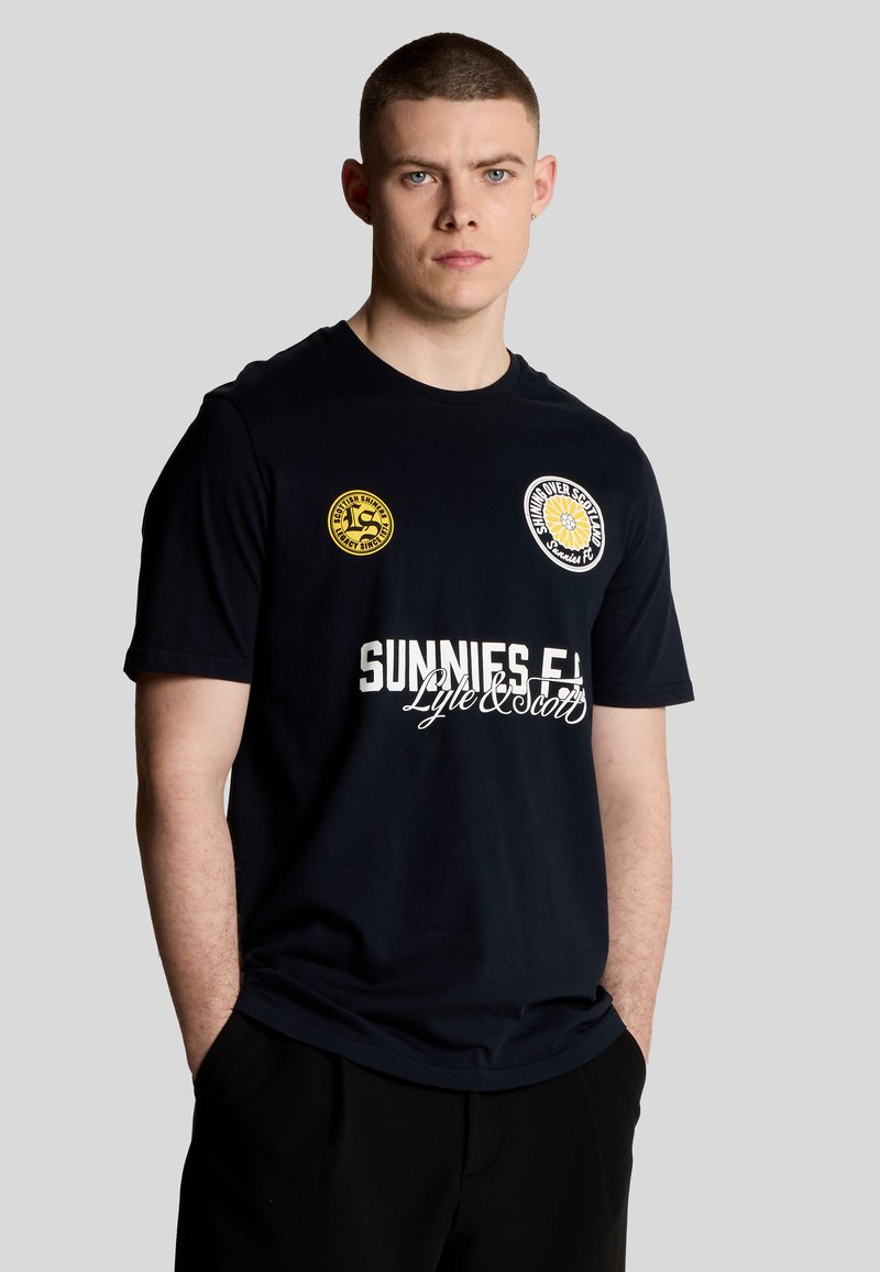 Marinblå t-shirt med kort ärm, prydd med vit text som säger "SUNNIES F.C." och två runda märken i gult och vitt på bröstområdet.