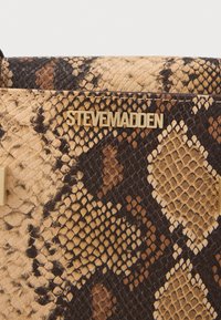 Textūruota dirbtinė gyvatės odos rankinė, smėlio ir rudos spalvos, su auksiniu "STEVE MADDEN" logotipu ir geometriniais akcentais.