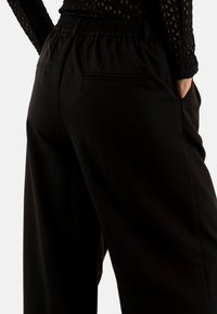 Pantaloni neri a vita alta con fascia elastica, dotati di tasche laterali e una consistenza liscia, mostrati da un'angolazione posteriore.