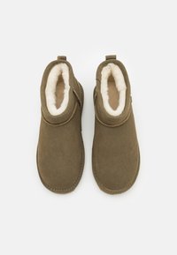 Stivaletti alla caviglia in pelle scamosciata verde oliva con morbida fodera in pile beige, punte arrotondate e linguette posteriori per tirare. Texture liscia con dettagli di cuciture visibili.