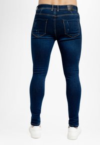 Jeans skinny de mezclilla azul oscuro con una textura suave, que cuentan con bolsillos traseros y una etiqueta de cuero en la cintura. Zapatillas blancas visibles.
