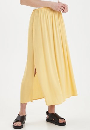 Personne portant une jupe maxi jaune clair avec une fente latérale et des sandales noires à bout ouvert, debout sur un fond blanc.