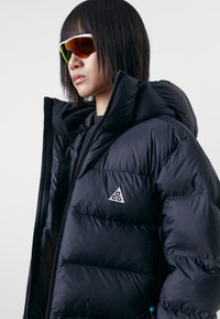 Black puffer jacket with a high collar, featuring a logo patch and zippered pockets. Smooth, shiny texture, and durable fabric.