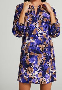 Kobieta w sukience do kolan w niebieskie, brązowe, czarne i białe abstrakcyjne plamy, poprawiająca złotą naszyjnik.