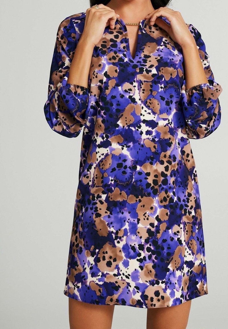 Kobieta w sukience do kolan w niebieskie, brązowe, czarne i białe abstrakcyjne plamy, poprawiająca złotą naszyjnik.