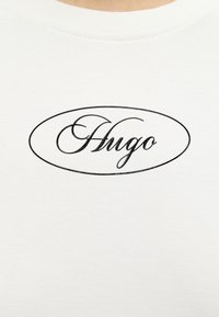 Λευκό ύφασμα με τη λέξη «Hugo» σε μαύρη καλλιγραφική γραφή, περικλεισμένη σε ένα απλό οβάλ περίγραμμα.