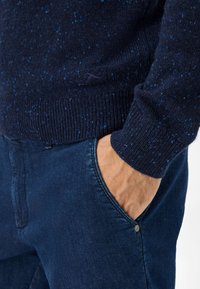 Marineblauer gestrickter Pullover mit strukturierten Sprenkeln, gerippten Bündchen und taillierter Passform. Kombiniert mit dunkelblauem Denim, der seitliche Taschendetails zeigt.
