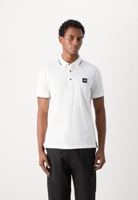 Polo shirt bianco con dettagli neri, con due bottoni e una piccola etichetta nera sul petto. Maniche corte e vestibilità rilassata.
