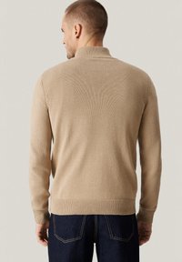 Beżowy sweter z dzianiny z wysokim kołnierzem i prążkowanym wykończeniem, z długimi rękawami i gładką fakturą. Noszony z ciemnymi dżinsami.