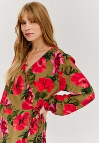 Kwiatowa sukienka w stylu wrap z zielonym tłem oraz dużymi czerwonymi i różowymi kwiatami, długimi bufiastymi rękawami oraz elastycznym pasem z troczkami umożliwiającym regulację dopasowania.