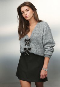 Grauer Strickpullover mit zwei schwarzen Satin-Schleifen und weißen Verzierungen, kombiniert mit einem schwarzen Rock. Silberne Armbanduhr am Handgelenk. Glatter Hintergrund.