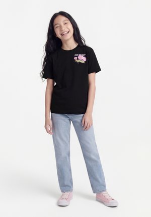 Χαμογελαστή κοπέλα με μακριά μαύρα μαλλιά φοράει μαύρο φλοράλ μπλουζάκι Off-White, ανοιχτό μπλε τζιν και ροζ αθλητικά παπούτσια, στέκεται μπροστά σε λευκό φόντο.