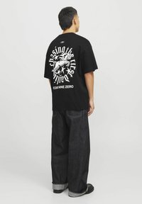 Sort oversized t-shirt med hvid grafisk print, der viser en figur, stjerner og tekst på ryggen. Kombineret med mørk denimjeans.