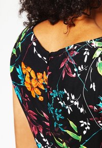 Femme portant une robe noire avec des motifs floraux colorés, montrant l'encolure dans le dos et des cheveux bouclés.
