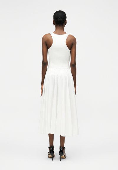 Γυναίκα με την πλάτη γυρισμένη φοράει άσπρο ριμπ αμάνικο τοπ, λευκή πλισέ midi φούστα και μαύρα ψηλοτάκουνα σανδάλια σε μονόχρωμο φόντο.