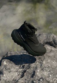 Black hiking shoe with a high ankle collar, textured synthetic upper, and a chunky rubber sole, shown mid-air above a rocky surface.