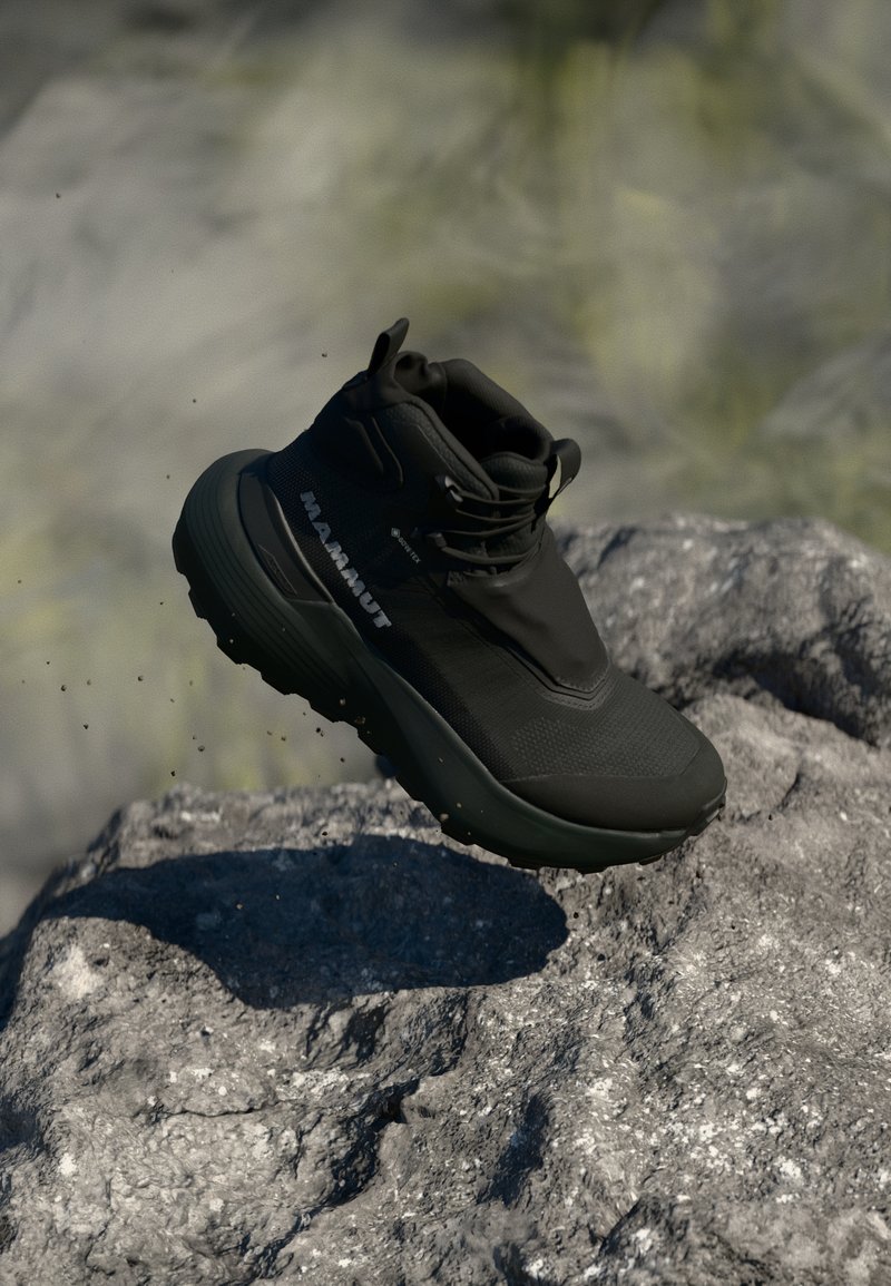 Black hiking shoe with a high ankle collar, textured synthetic upper, and a chunky rubber sole, shown mid-air above a rocky surface.