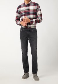 Camicia a quadri rossa, bianca e blu navy con colletto button-down; jeans neri; scarpe in suede grigie con dettagli bianchi. Il modello si trova in posa casual.