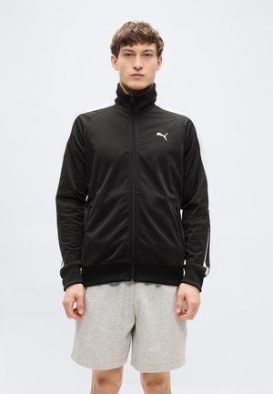 Zwarte jas met ritssluiting, gemaakt van gladde stof met geribbelde manchetten en kraag. Voorzien van witte zijstrepen en een klein wit logo op de borst.