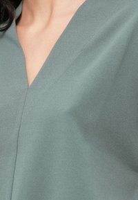Απαλή, υφασμάτινη υφή σε απαλό πράσινο, με σχέδιο V-neck και διακριτική πτυχοποίηση, με ορατές ραφές που ενισχύουν το σχήμα.