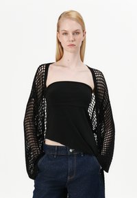 Cardigã de malha preta com trama aberta, usado sobre um top preto sem alças, combinado com calções de ganga de cintura alta azuis. Design simples, sem padrões.