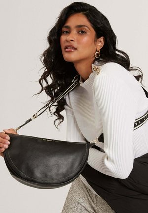 Femme aux longs cheveux bouclés portant un pull blanc côtelé et des boucles d'oreilles en anneau doré, tenant un sac à main en cuir noir en forme de demi-lune.