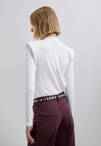 Wit geribd shirt met lange mouwen en schoudervullingen, gecombineerd met bordeauxrode broek met een zwarte riem met studs. Achteraanzicht toont de pasvorm van de outfit.