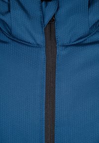 Blå jacka med en hexagonal mönstrad textur, med en central svart dragkedja och en huva vid halslinningen. Slät tygfinish.