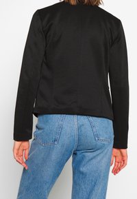 Femme portant un blazer noir cintré et un jean taille haute bleu clair, vue de dos sur un fond blanc uni.