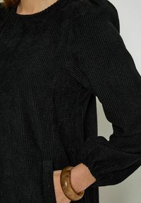 Black corduroy dress featuring a textured finish, round neckline, and puffed sleeves. A light brown bracelet complements the outfit.