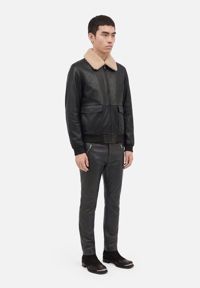 Homme debout portant une veste en cuir noire avec un col en fourrure beige, un pantalon en cuir noir et des bottes noires sur un fond uni.