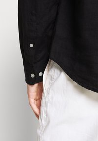 Černá strukturovaná lněná košile s viditelnými knoflíky na manžetách, nošena přes světlé kalhoty. Zaměření na detail rukávu a design manžety.